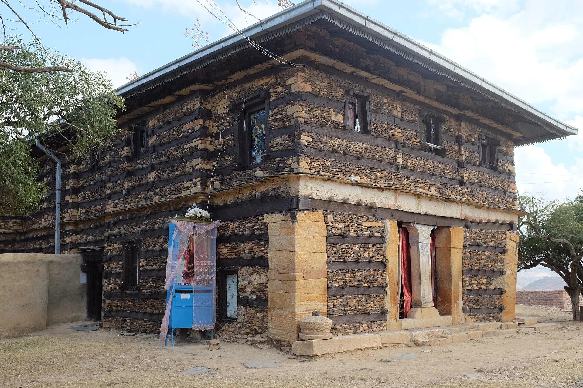 Colour three-quarter photograph of the Däbrä Dammo church from the entrance corner, showing two storeys of banded stone and timber walling, large dressed sandstone quoins, deep overhanging eaves with a pierced metal valance, and small upper windows