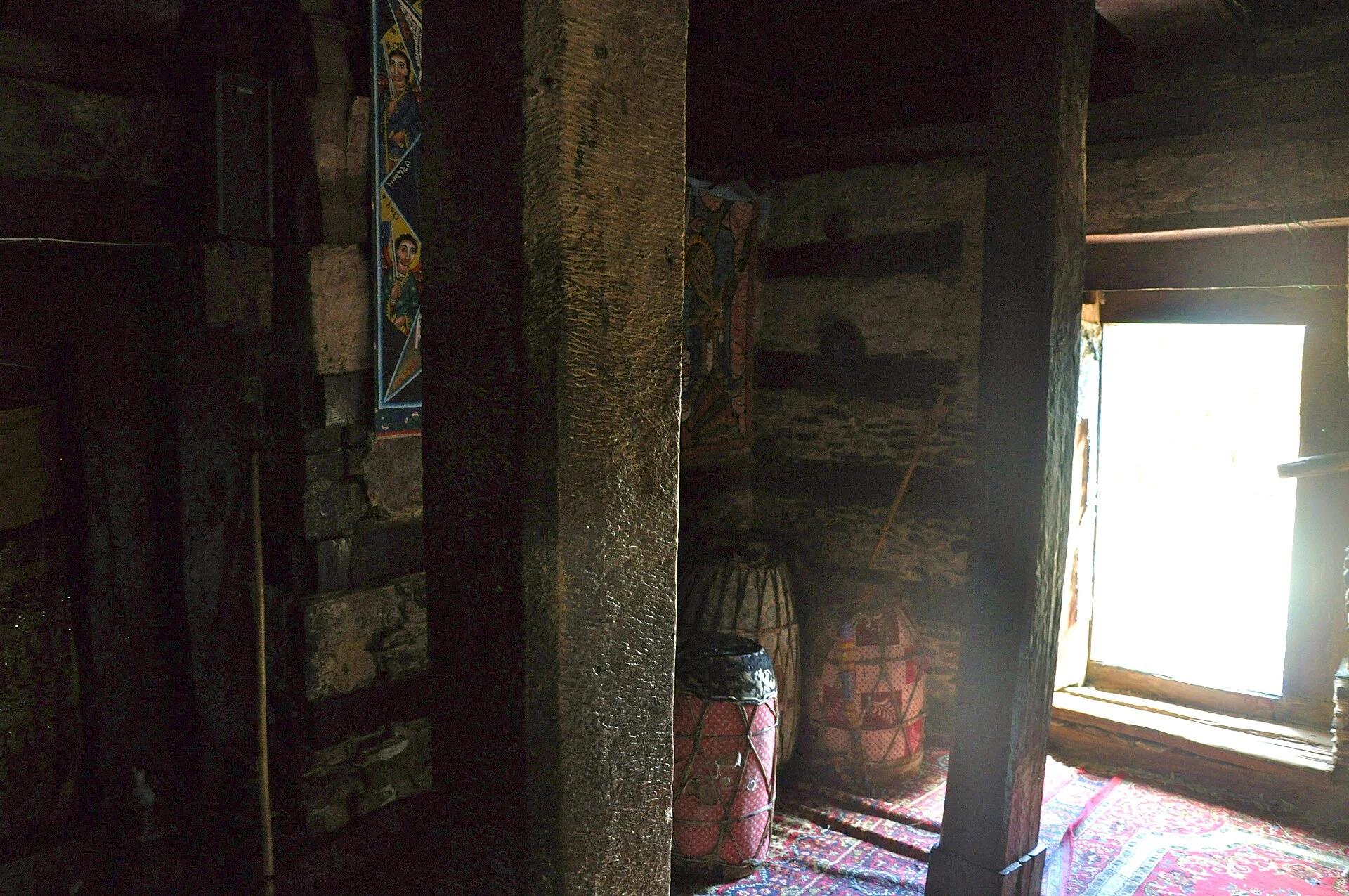 Dim colour interior photograph of the church: a pale tooled stone pier and a near-black timber post stand against rubble walls in deep shadow, while a deep timber-lined window on the right burns out to white daylight