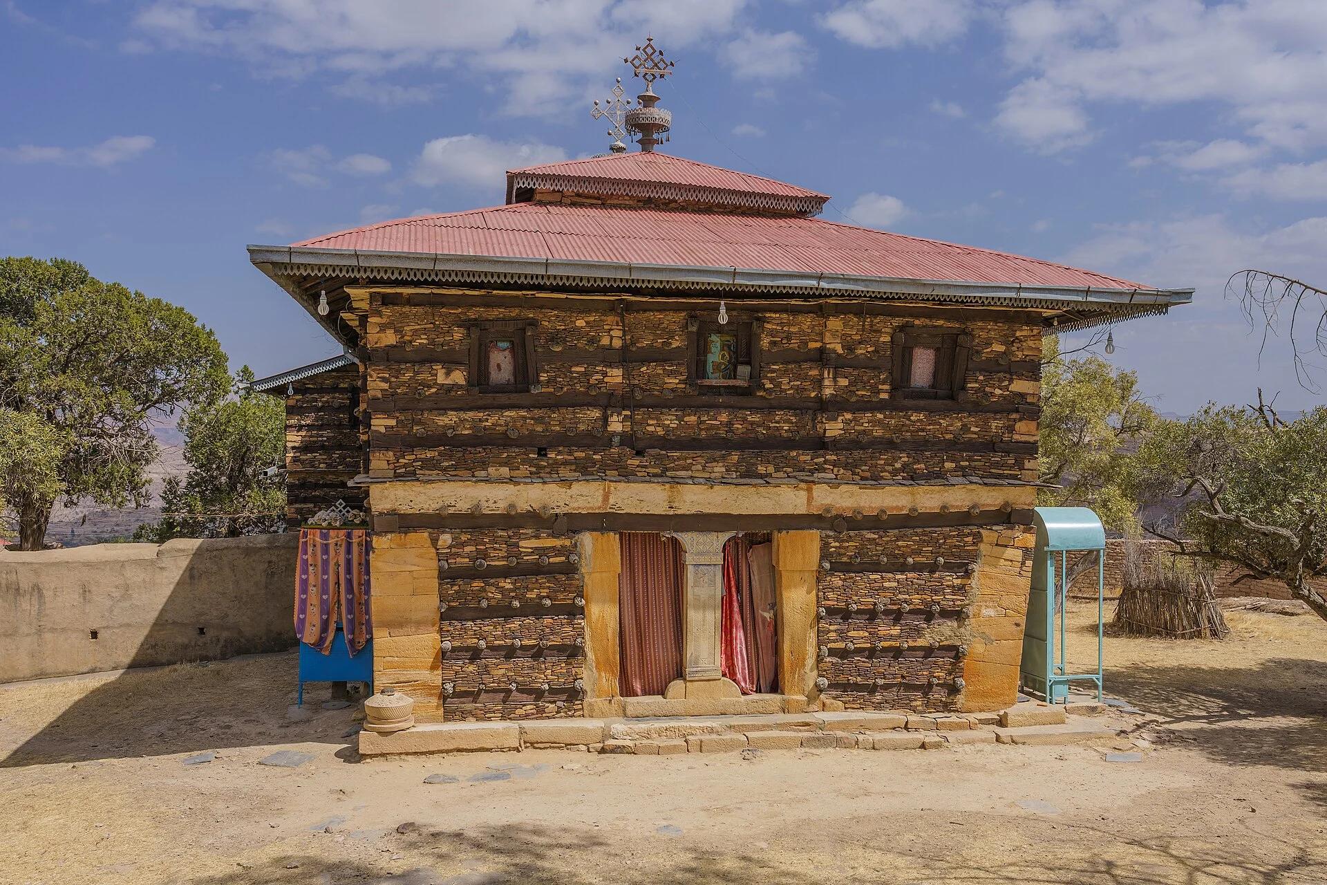 Colour photograph of the west front of the Däbrä Dammo church: banded walls of thin honey-coloured stone slabs and dark timber sills studded with pale round beam ends, ochre dressed-stone door piers, three small painted windows above, and a red corrugated roof with a crowned cross finial