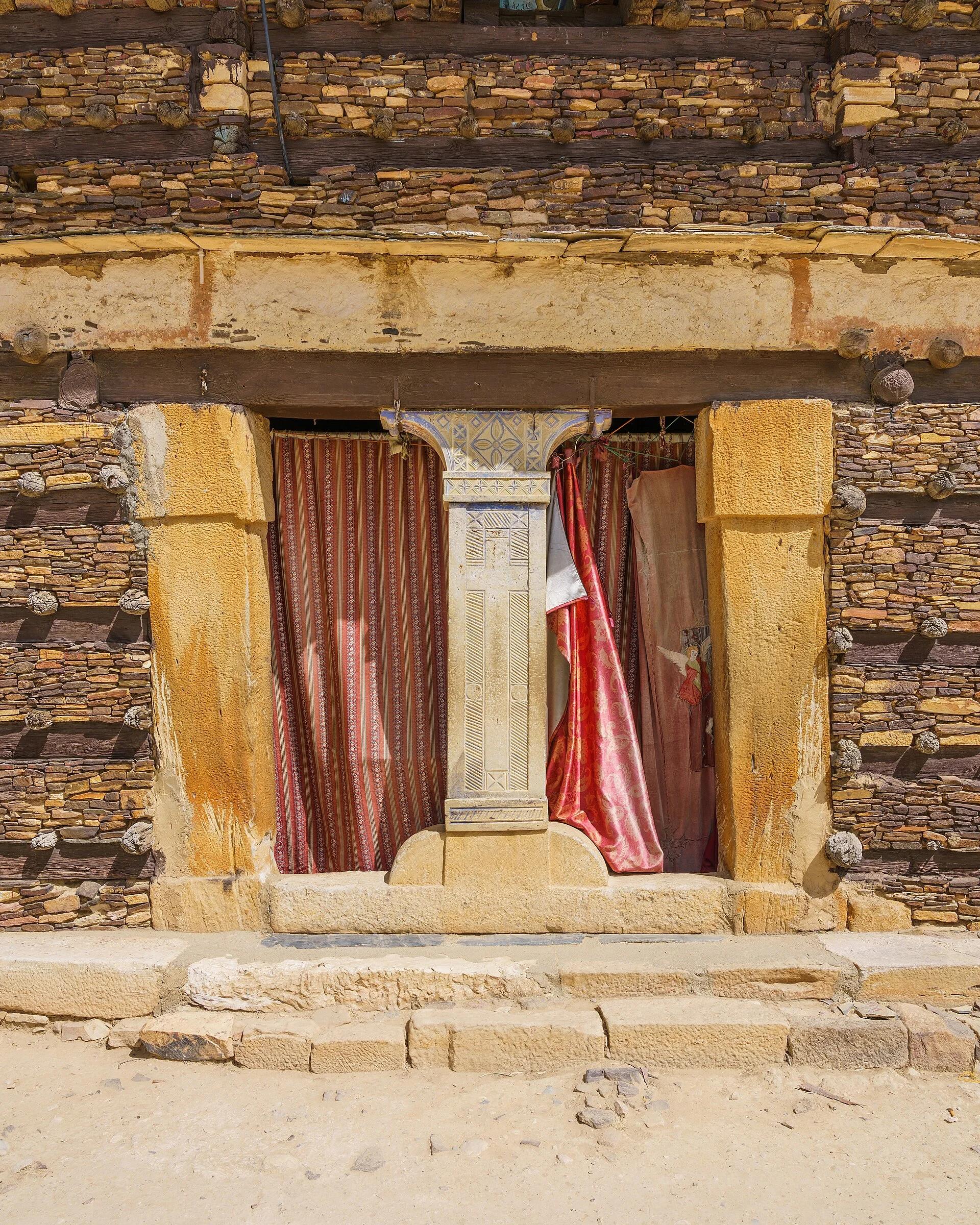 Close colour photograph of the church entrance: massive ochre sandstone jambs, a carved pale stone mullion incised with a cross, a heavy timber lintel, and the surrounding wall of thin stone slabs banded by dark timbers with projecting pale beam ends