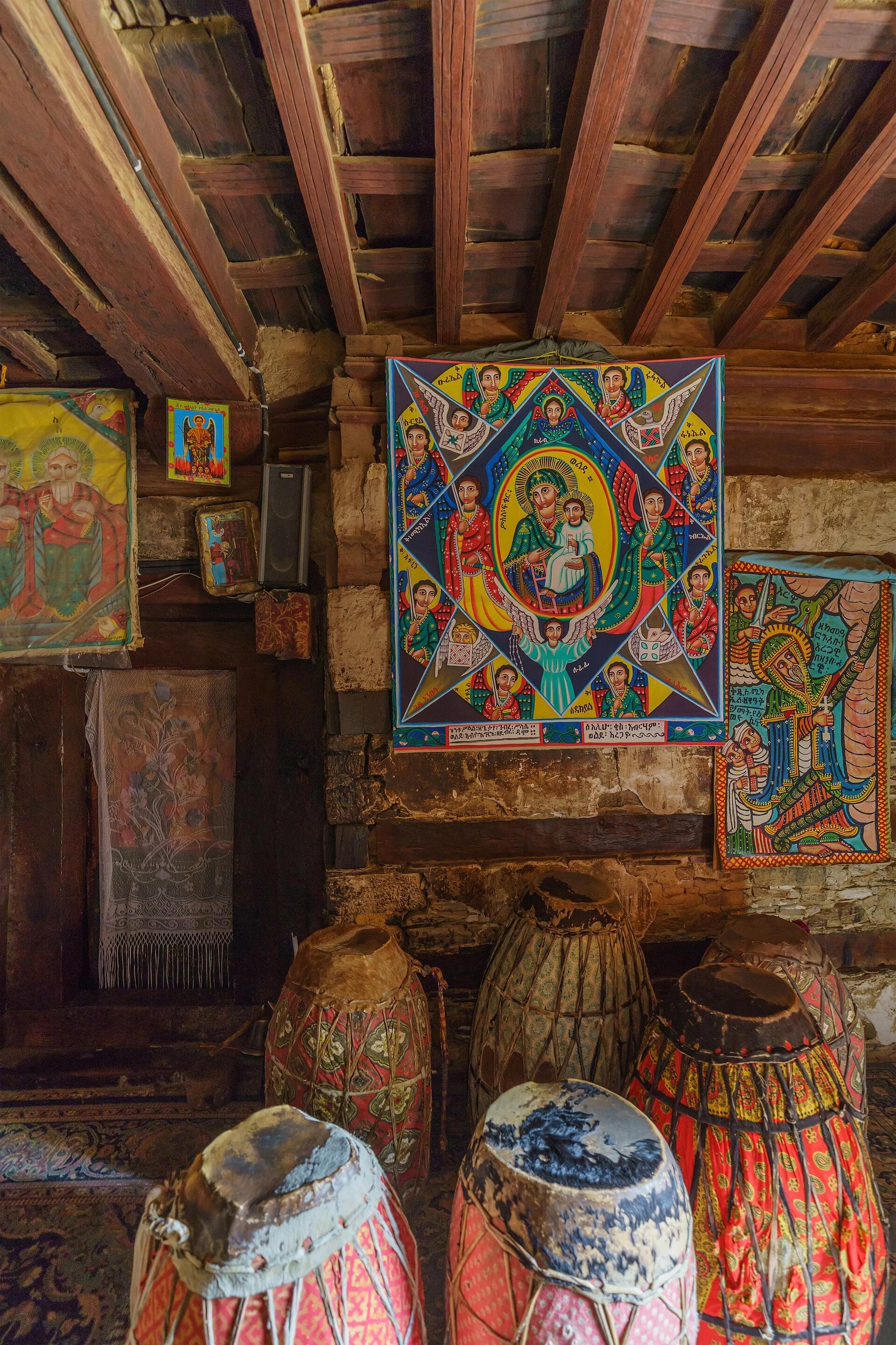 Colour interior photograph of a ceiling of closely spaced reddish-brown timber joists carrying boards, above a wall of pale stone banded with dark timber, hung with painted icon panels, with large drums standing below