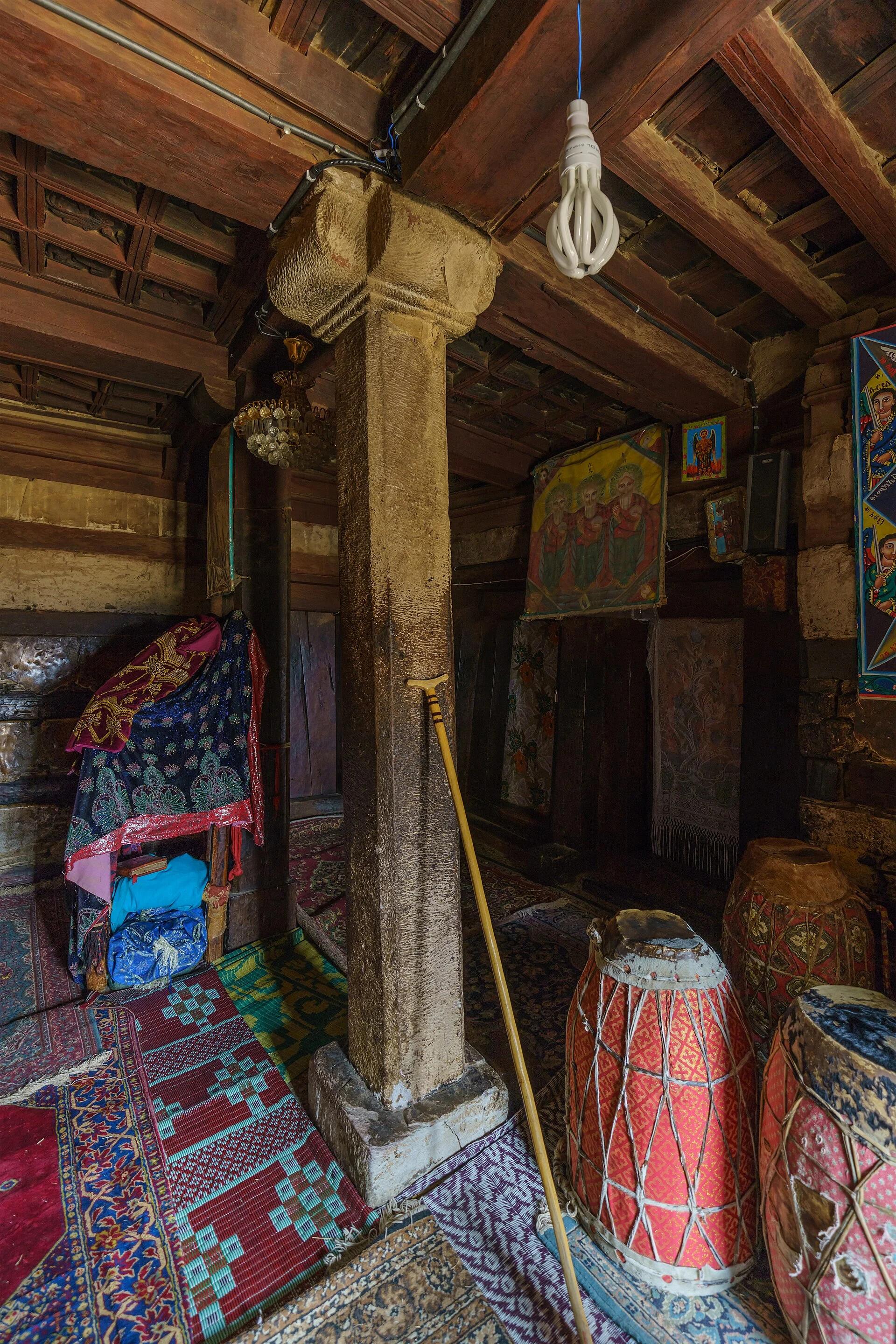 Colour interior photograph of a square stone monolith in the church, its shaft covered in vertical tool marks, spreading through a moulded impost to a broad capital that carries dark timber beams and a carved coffered ceiling; drums, carpets and painted icons stand around it