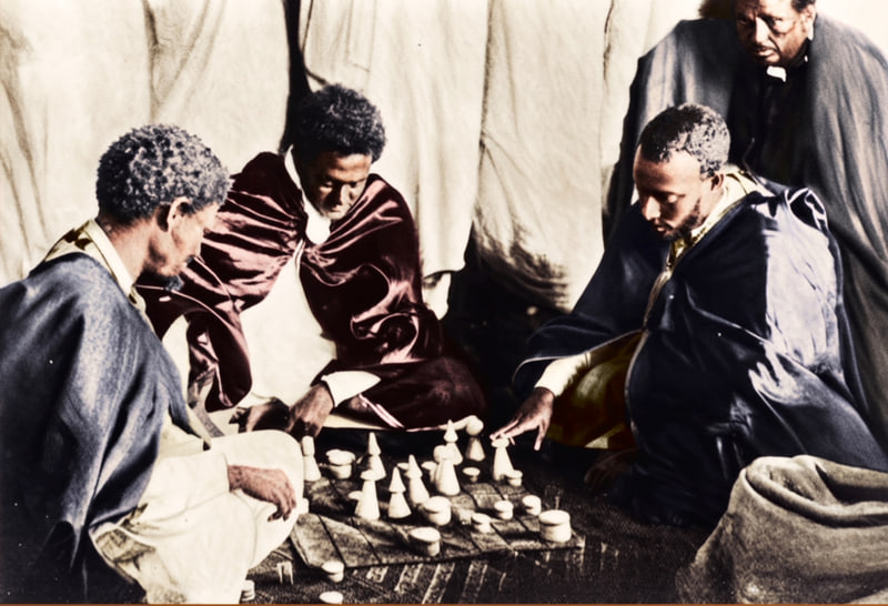 Four men seated on the ground around a ruled cloth board of turned playing pieces inside a tent, in an early twentieth-century photograph, colourised.